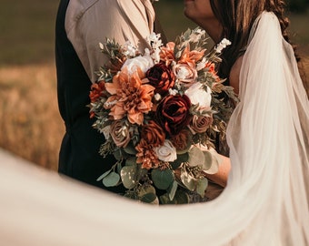 May include: A bride and groom are embracing, their foreheads touching. The bride is wearing a white veil and holding a bouquet of burgundy, peach, and white flowers. The groom is wearing a brown shirt and a black vest.