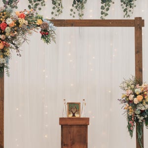 K&ouml;nnte beinhalten: Eine h&ouml;lzerne Hochzeitsbogen mit farbenfrohen Blumengestecken. Der Bogen ist mit Blumen in Orange-, Gelb- und Wei&szlig;t&ouml;nen geschm&uuml;ckt. Ein kleines Holzpodest mit einem gerahmten Bild und zwei Glasflaschen befindet sich vor dem Bogen.