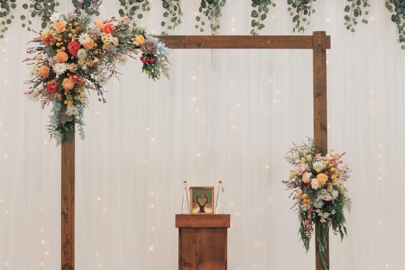 K&ouml;nnte beinhalten: Eine h&ouml;lzerne Hochzeitsbogen mit farbenfrohen Blumengestecken. Der Bogen ist mit Blumen in Orange-, Gelb- und Wei&szlig;t&ouml;nen geschm&uuml;ckt. Ein kleines Holzpodest mit einem gerahmten Bild und zwei Glasflaschen befindet sich vor dem Bogen.