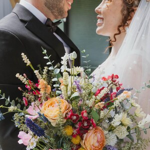 K&ouml;nnte beinhalten: Ein lebendiger Blumenstrau&szlig; mit pfirsichfarbenen, rosa, roten und wei&szlig;en Blumen mit gr&uuml;nem Laub. Der Strau&szlig; wird vor einer Person in einem schwarzen Smoking und einer Person in einem wei&szlig;en Kleid und Schleier gehalten.