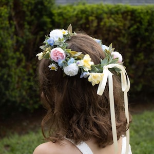 K&ouml;nnte beinhalten: Ein Blumenkranz mit rosa Rosen, wei&szlig;en G&auml;nsebl&uuml;mchen und gelben Bl&uuml;ten, erg&auml;nzt durch gr&uuml;ne Bl&auml;tter. Cremefarbene B&auml;nder fallen nach hinten. Die Person tr&auml;gt ein wei&szlig;es schulterfreies Oberteil. Dekoratives Haaraccessoire.