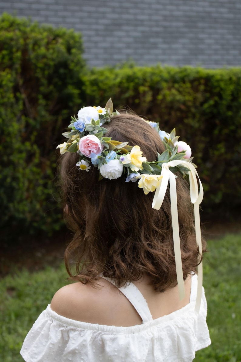 K&ouml;nnte beinhalten: Ein Blumenkranz mit rosa Rosen, wei&szlig;en G&auml;nsebl&uuml;mchen und gelben Bl&uuml;ten, erg&auml;nzt durch gr&uuml;ne Bl&auml;tter. Cremefarbene B&auml;nder fallen nach hinten. Die Person tr&auml;gt ein wei&szlig;es schulterfreies Oberteil. Dekoratives Haaraccessoire.