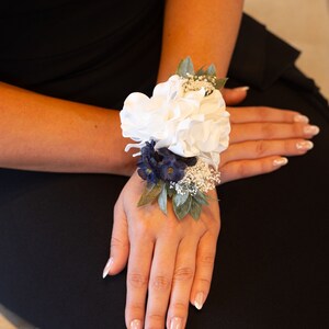 May include: A white flower wrist corsage with blue and white accents. The corsage is tied with a green ribbon and features a white flower in the centre.