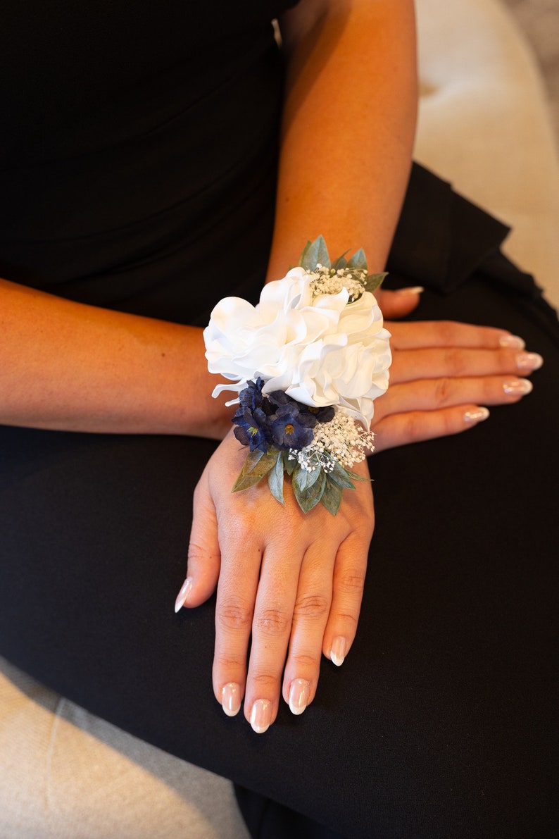 May include: A white flower wrist corsage with blue and white accents. The corsage is tied with a green ribbon and features a white flower in the centre.