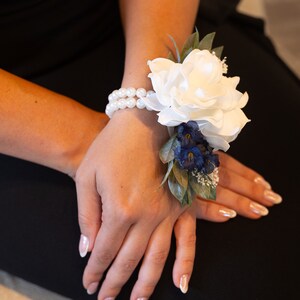 May include: A white flower wrist corsage with blue flowers and green leaves. The corsage is tied with a white pearl bracelet.