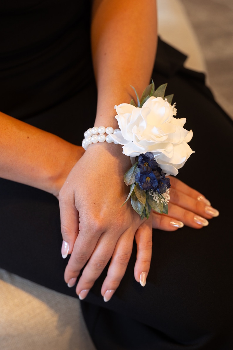 May include: A white flower wrist corsage with blue flowers and green leaves. The corsage is tied with a white pearl bracelet.