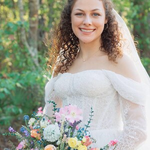 K&ouml;nnte beinhalten: Eine Braut in einem wei&szlig;en schulterfreien Brautkleid aus Spitze h&auml;lt einen bunten Blumenstrau&szlig;. Der Strau&szlig; enth&auml;lt rosa, blaue, gelbe und orangefarbene Bl&uuml;ten. Die Braut l&auml;chelt, und ein Schleier ist &uuml;ber ihrem Kopf drapiert.