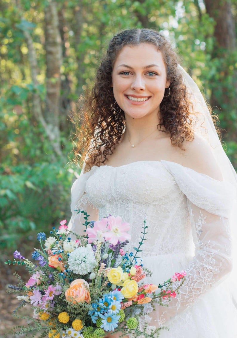 K&ouml;nnte beinhalten: Eine Braut in einem wei&szlig;en schulterfreien Brautkleid aus Spitze h&auml;lt einen bunten Blumenstrau&szlig;. Der Strau&szlig; enth&auml;lt rosa, blaue, gelbe und orangefarbene Bl&uuml;ten. Die Braut l&auml;chelt, und ein Schleier ist &uuml;ber ihrem Kopf drapiert.