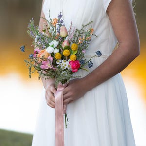 K&ouml;nnte beinhalten: Ein farbenfroher Blumenstrau&szlig; mit rosa, gelben, wei&szlig;en und orangen Bl&uuml;ten, gebunden mit einem rosa Band. Der Strau&szlig; wird vor einem wei&szlig;en Kleid gehalten.