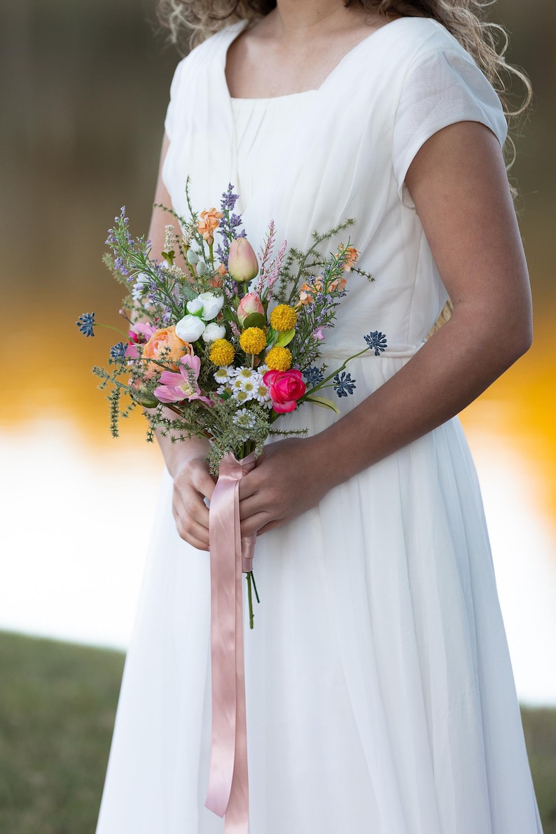 K&ouml;nnte beinhalten: Ein farbenfroher Blumenstrau&szlig; mit rosa, gelben, wei&szlig;en und orangen Bl&uuml;ten, gebunden mit einem rosa Band. Der Strau&szlig; wird vor einem wei&szlig;en Kleid gehalten.