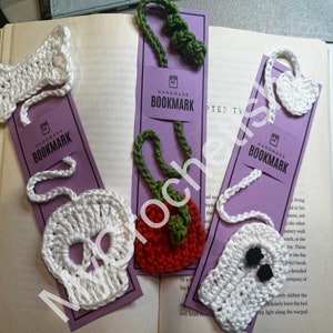 Può includere: Tre segnalibri fatti a mano all'uncinetto con retro in cartoncino viola. I segnalibri presentano un osso bianco, una mela rossa e verde e un teschio bianco. Ogni segnalibro ha il testo "Segnalibro fatto a mano" stampato sul cartoncino.