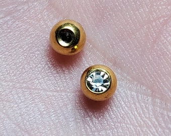 Op de afbeelding: Twee gouden lichaamsieraden, een met een heldere strasssteen en een zonder.