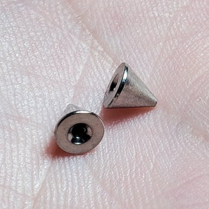 May include: Two silver cone-shaped body jewelry pieces. One piece is a flat base with a screw-on top. The other piece is a cone with a rounded tip.