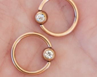 Op de afbeelding: Twee gouden ronde ringen met een heldere edelsteen ingebed in elk. De ringen zijn ontworpen voor body piercing.