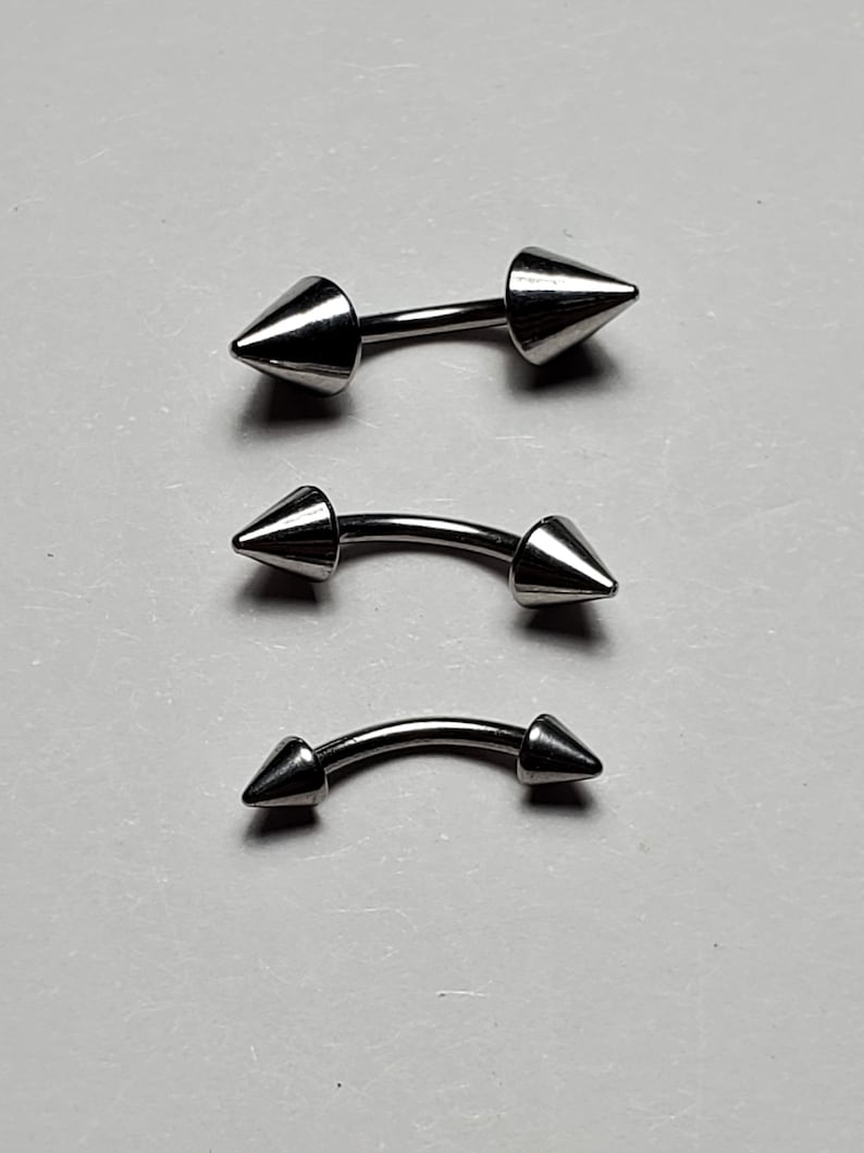 May include: Three silver barbell body jewelry pieces with pointed ends. The barbells are different sizes.