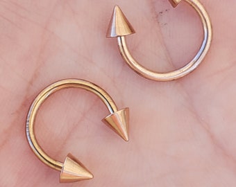 Op de afbeelding: Twee gouden ronde barbell piercings met puntige uiteinden. De piercings liggen op een lichtroze oppervlak.