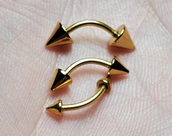 Op de afbeelding: Drie gouden lichaamssieraden met puntige uiteinden. De sieraden zijn in de vorm van een gebogen halter.