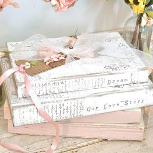 May include: A stack of three books with white covers featuring grey script text. The top book is tied with a sheer white ribbon and a pink ribbon, adorned with small pink flowers and a brown tag. The spines are labeled with the words Dream and Our Love Story.