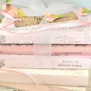 May include: A stack of decorative books in shades of pink and white, featuring butterfly and script designs. The books are tied with a pink ribbon and decorated with faux flowers and a tag. The text on the books includes phrases like "have faith always" and "there is always hope."