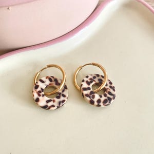 Puede incluir: Un par de pendientes de aro dorados con un dije circular con estampado de leopardo. Los pendientes se muestran sobre un fondo claro, resaltando el oro y las manchas marrones y crema del dije.