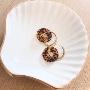 Puede incluir: Un par de pendientes de aro dorados con colgantes circulares con estampado de carey descansan sobre una fuente blanca en forma de concha con borde dorado. Los pendientes son un accesorio elegante.