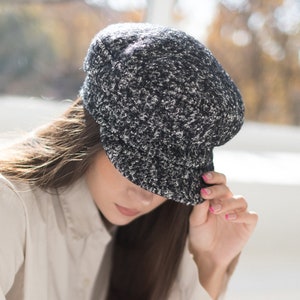 May include: A woman wearing a white button-down shirt and black pants is holding a black and white tweed newsboy cap.