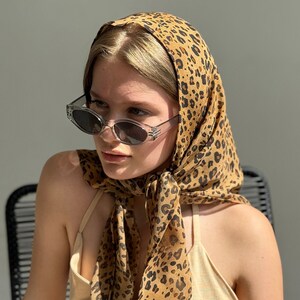 May include: A woman wearing a brown and black leopard print headscarf tied in a knot at the back of her neck. She is wearing clear framed sunglasses and a light brown sleeveless dress.