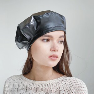 May include: A black leather beret with a smooth, shiny finish. The beret is worn by a person with long brown hair.