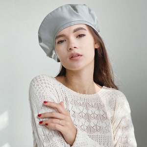May include: A woman wearing a light gray beret and a white crocheted sweater. She is looking at the camera with her arms crossed.