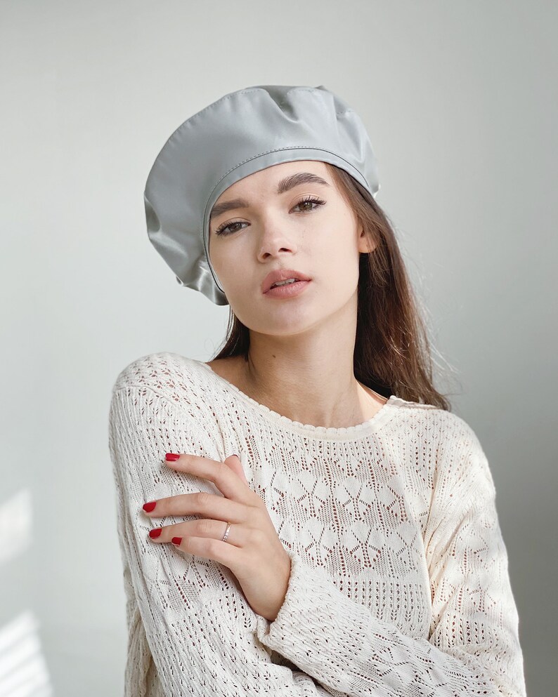 May include: A woman wearing a light gray beret and a white crocheted sweater. She is looking at the camera with her arms crossed.