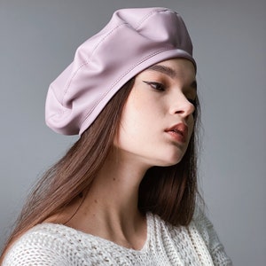 May include: A woman wearing a light pink leather beret and a white knitted sweater.