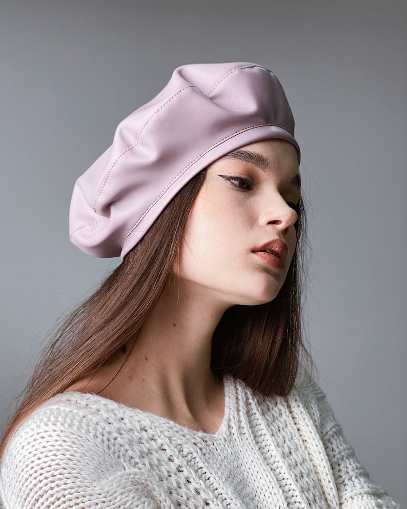 May include: A woman wearing a light pink leather beret and a white knitted sweater.