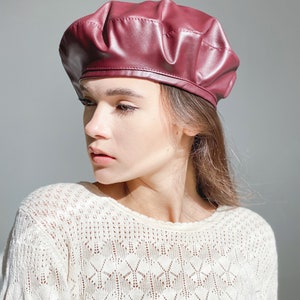 May include: A woman wearing a maroon leather beret and a white crocheted sweater.