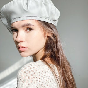 May include: A light gray beret made of faux leather. The beret has a slightly wrinkled texture and a simple design.