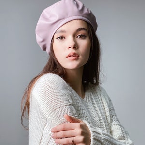 May include: A woman wearing a light pink beret and a white knitted sweater. She is looking at the camera with a neutral expression.