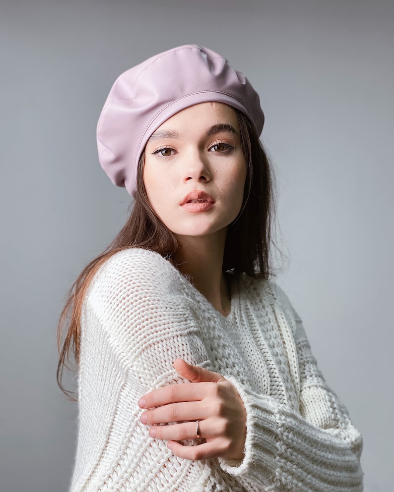May include: A woman wearing a light pink beret and a white knitted sweater. She is looking at the camera with a neutral expression.