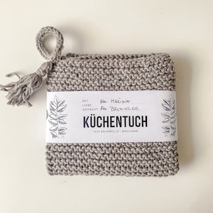 Könnte beinhalten: Ein graues, gestricktes Geschirrtuch mit einem Quasten. Das Tuch hat ein Etikett mit der Aufschrift "Mit Liebe Gemacht" und "Küchen Tuch" mit dem Text "100% Baumwolle - Waschbar". Das Etikett enthält auch den Text "Von: Mariwa" und "Für: Brun Hilde".
