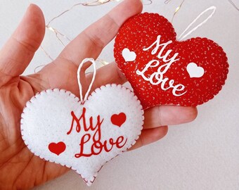 Könnte beinhalten: Zwei Filz-Herzornamente mit dem Text "My Love" in roten Buchstaben. Ein Herz ist rot mit weißen Glitzer und das andere ist weiß mit roten Glitzer.