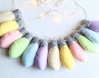 May include: A garland of felt light bulbs in pastel colors, including light pink, yellow, light blue, and lavender. The bulbs are strung together with gray felt and a white string.