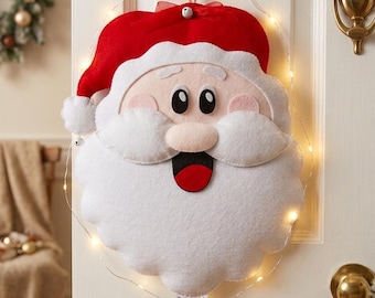 May include: A felt Santa Claus face wall hanging featuring a red hat with a white pom-pom, a large white beard, a pink nose, and a wide open mouth. The decoration is outlined with a thin wire string of warm white LED lights and hangs from a hook.