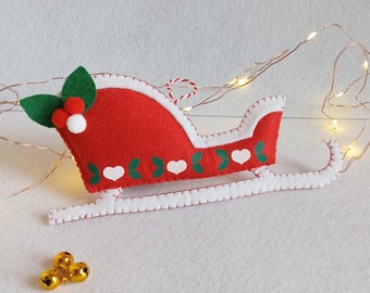May include: Red felt Christmas sleigh ornament with white hearts and green holly leaves. The sleigh has a white felt trim and a red and white string for hanging.