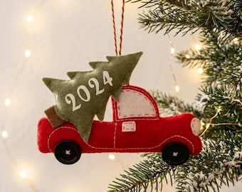 May include: A felt Christmas ornament shaped like a red truck carrying a green Christmas tree with the year 2024 printed in white. The truck features black button wheels and white stitching details, suspended by a red and white twine string.