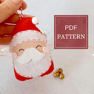 Könnte beinhalten: Handgefertigter Santa Claus-Ornament aus Filz mit rotem Hut und Anzug, weißem Bart und rosigen Wangen. Das Ornament wird von einer Hand gehalten und hat eine rot-weiße Schnur zum Aufhängen. Der Text "PDF PATTERN" ist in einem roten Feld zu sehen.
