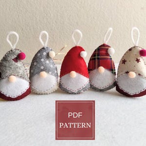 Può includere: Cinque ornamenti di gnomi fatti a mano con barbe bianche, nasi beige e cappelli rossi, grigi e fantasia. Ogni ornamento ha un anello bianco per appenderli. L'immagine include una scatola rossa con il testo "PDF PATTERN".