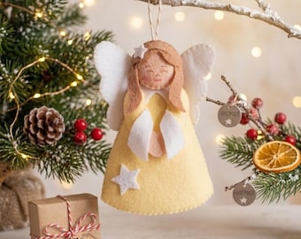 Könnte beinhalten: Handgefertigter Weihnachtsbaumschmuck aus Filz, der einen Engel mit gelbem Kleid, weißen Flügeln und einem Stern darstellt. Der Engel hängt an einem Ast, mit einer Geschenkbox und einem Weihnachtsbaum im Hintergrund. Das Etikett auf der Geschenkbox lautet "Minasfactory".