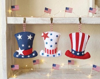 Könnte beinhalten: Drei Uncle Sam-Hüte aus Filz in Rot, Weiß und Blau, verziert mit Sternen und Streifen. Jeder Hut hängt an einer rot-weißen Schnur. Kleine amerikanische Flaggen und Lichterketten schmücken das Regal.