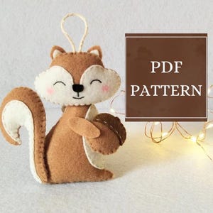 Puede incluir: Adorno de ardilla de fieltro hecho a mano en tonos marrón, crema y blanco, sosteniendo una bellota de fieltro. La ardilla tiene una cara sonriente con mejillas rosadas y un lazo para colgar. La imagen incluye el texto "PDF PATTERN".