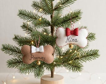 May include: Two bone-shaped felt ornaments hanging on a small evergreen tree. One ornament is tan with a white bow and the name Rooney embroidered in black. The other is white with a red glittery bow and the name Kira embroidered in black.