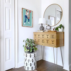 Puede incluir: Un armario de madera con cajones y patas de horquilla, un espejo redondo con marco dorado, una l&aacute;mpara blanca con pantalla gris, una planta en maceta y un taburete con estampado blanco y negro.