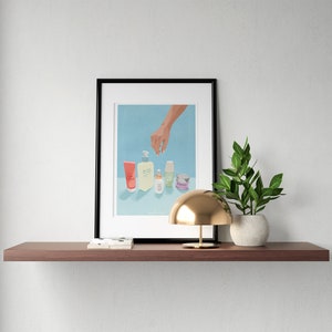 May include: A framed illustration of a hand reaching for a row of five different skincare products on a light blue background. The products include a pink tube, a clear bottle with a pump, a small clear bottle, a green bottle, and a small purple jar. The text "Daily Routine" is written below the products.